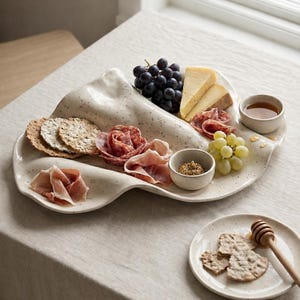May include: A speckled, off-white ceramic serving platter with a wavy design, filled with grapes, cheese, crackers, cured meats, and small bowls of condiments. A smaller plate with crackers and a honey dipper is also present.