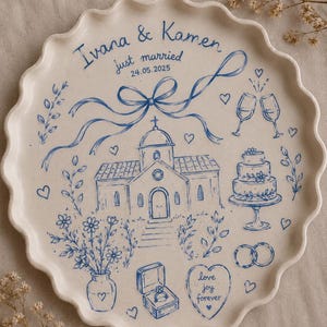 Puede incluir: Una fuente de cerámica blanca con borde festoneado, pintada a mano con ilustraciones azules. Presenta una iglesia, tarta de boda, anillos y el texto "Ivana & Kamen just married 24.05.2025".