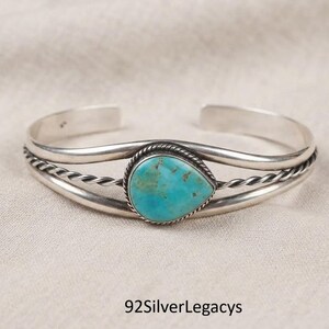 May include: A silver cuff bracelet featuring a central teardrop-shaped turquoise stone. The band consists of two smooth silver wires flanking a central twisted rope wire, with the turquoise stone set in a matching twisted rope silver bezel.
