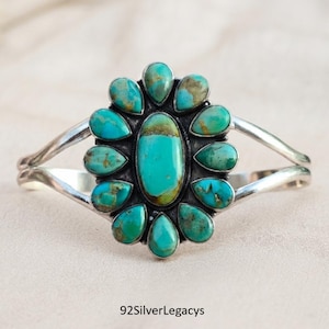 May include: A silver cuff bracelet featuring a large central oval turquoise stone surrounded by twelve smaller teardrop-shaped turquoise stones. The metal band splits into two arms on each side. The text 92SilverLegacys is visible at the bottom.