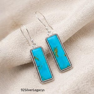 May include: A pair of drop earrings featuring rectangular turquoise stones set in silver frames with a twisted rope border. The earrings have silver hook wires and display natural brown matrix patterns within the bright blue stones.