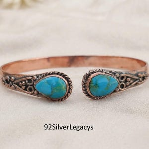 May include: A copper cuff bracelet featuring two teardrop-shaped turquoise stones at the open ends. The metal band displays intricate embossed scrollwork and bead detailing surrounding each stone. The text 92SilverLegacys is visible.