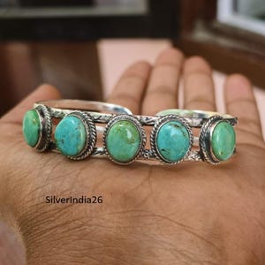 May include: A silver cuff bracelet featuring five oval-shaped turquoise stones set in rope-textured bezels. The metal band displays intricate engraved detailing along the sides. The text SilverIndia26 is visible on the lower left side.