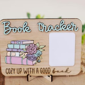 Puede incluir: Rastreador de libros de madera con las palabras "Book tracker" en escritura azul claro. El diseño incluye una pila de libros con detalles florales y un espacio en blanco. También se incluye la frase "Cozy up with a good book".