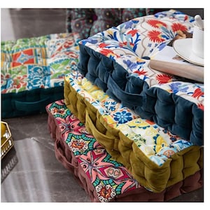 May include: A stack of colorful floor cushions with floral and geometric patterns. The cushions have a tufted design with velvet sides and carrying handles. The color palette includes red, blue, yellow, and green.