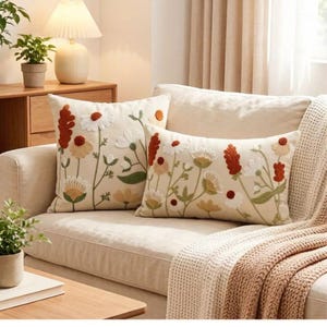 May include: Two cream-coloured throw pillows featuring embroidered floral designs in shades of rust orange, sage green, and white. The square and rectangular pillows display various wildflowers and stems, showcasing textured stitching and botanical patterns.