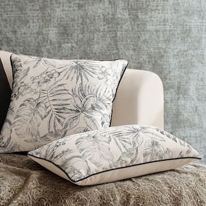 Può includere: Due cuscini quadrati con una base in tessuto color crema e una stampa botanica grigio scuro di foglie e fiori tropicali. Ogni cuscino è rifinito con un sottile bordino nero, che conferisce un contorno definito al motivo floreale.