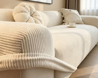 Op de afbeelding: Een crèmekleurige bank met een geribbelde, corduroy-achtige textuur, voorzien van een rond, getuft sierkussen en een vierkant kussen. Een klein wit keramieken kopje met schoteltje staat op het zitkussen. Een ingelijst bord toont Sfontara.