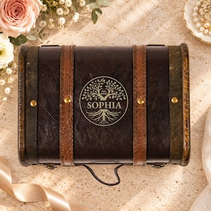 May include: A dark brown rectangular storage box with two vertical brown leather-like straps and brass-colored studs. The center features a circular engraving of a tree with the name SOPHIA and the text est. 2022 printed below it.