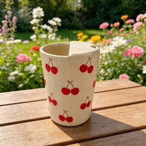 May include: A cream-colored ceramic mug with a handle and a cherry pattern. The mug is cylindrical with a slightly flared top and is decorated with red cherries. The mug is sitting on a wooden surface.
