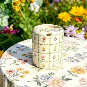 Puede incluir: Un recipiente cilíndrico de cerámica con tapa, decorado con un patrón de cuadrícula y pequeñas flores coloridas. El recipiente está sobre un mantel floral, con un fondo borroso de flores coloridas.