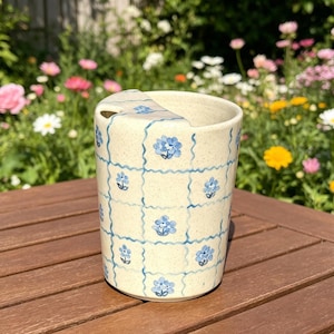 May include: A ceramic mug with a lid, cream-coloured with a blue floral pattern. The design features a grid of blue lines and blue flower motifs. The mug is on a wooden surface.