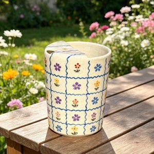 May include: A cream-colored ceramic mug with a handle, decorated with a grid pattern of blue lines and colorful flowers. The flowers are in shades of purple, yellow, and orange. The mug is sitting on a wooden surface.