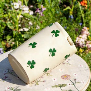 May include: Cream-colored ceramic planter with green shamrock designs. The cylindrical planter has a rectangular opening and a small handle. The planter is resting on a floral patterned surface, with a blurred background of greenery.