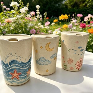 May include: Three ceramic tumblers with unique designs. One features blue waves and red starfish, another has a moon and stars, and the third showcases fish, coral, and seashells. Each tumbler is beige with a speckled texture.
