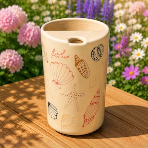May include: A cylindrical ceramic container with a cream-colored base featuring various printed illustrations of seashells, a starfish, and small botanical sprigs in shades of red, orange, and black. The top rim has a semi-circular cutout section.
