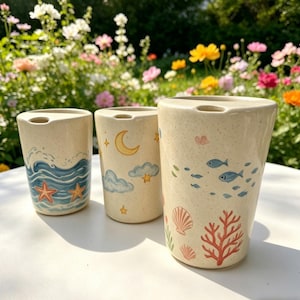 Puede incluir: Tres vasos de cerámica con diseños pintados a mano. Uno presenta olas y estrellas de mar, otro una luna y nubes, y el tercero peces, corales y conchas marinas. Cada vaso tiene un color beige moteado neutro.