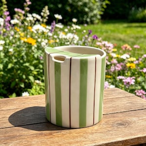 Puede incluir: Un recipiente de cerámica con un patrón de rayas verdes y crema. El recipiente tiene un asa curva y una pequeña abertura. El fondo presenta una superficie de madera y una vista borrosa de un jardín con flores coloridas.