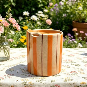 Puede incluir: Un recipiente cilíndrico de cerámica con rayas verticales naranjas. El recipiente tiene un asa y una tapa de madera. El fondo presenta un mantel floral y un jardín con flores.