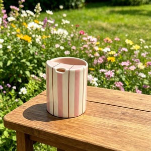 Puede incluir: Un recipiente cil&iacute;ndrico de cer&aacute;mica con rayas verticales rosas y marrones. Est&aacute; sobre una superficie de madera, con un jard&iacute;n lleno de flores coloridas en el fondo. El recipiente tiene una abertura en la parte superior.