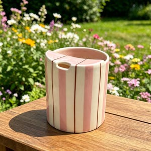 Puede incluir: Un jarr&oacute;n de cer&aacute;mica a rayas rosas y blancas. Tiene una abertura rectangular y una peque&ntilde;a ranura lateral. El jarr&oacute;n est&aacute; sobre una superficie de madera, con un jard&iacute;n al fondo.