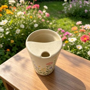 May include: A beige ceramic utensil holder with a floral design. The holder has a curved top and a small hole at the bottom. The floral design includes pink, yellow, and purple flowers with green stems and leaves.