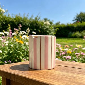 Puede incluir: Un jarr&oacute;n cil&iacute;ndrico de cer&aacute;mica con rayas verticales rosas y crema. El jarr&oacute;n tiene una peque&ntilde;a abertura en la parte superior y est&aacute; sobre una superficie de madera. El fondo presenta un jard&iacute;n con flores y vegetaci&oacute;n.