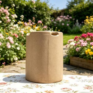 May include: A beige, cylindrical ceramic vase with two small, curved handles near the top. The vase has a textured surface and sits on a floral patterned tablecloth. The background features a garden with colorful flowers.