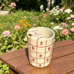 May include: A cream-colored ceramic mug with a lid, decorated with a red grid pattern and floral motifs. The mug sits on a wooden surface, with a garden background filled with flowers.