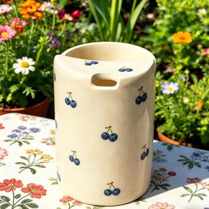 Puede incluir: Un soporte para utensilios de cerámica de color crema con forma rectangular redondeada. El soporte está decorado con pequeñas ilustraciones de cerezas azules. El fondo presenta un mantel floral y plantas en macetas con flores coloridas.