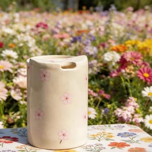 Op de afbeelding: Een crèmekleurige keramische vaas met een gebogen bovenkant en een handgreepachtige opening. De vaas is versierd met roze kersenbloesemmotieven. De achtergrond toont een veld met kleurrijke bloemen.