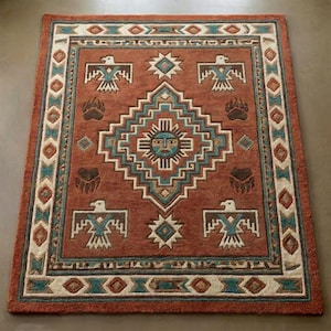 May include: A rectangular area rug with a Southwestern design. The rug features a rust-colored background with geometric patterns in teal, cream, and brown. The design includes stylized birds, bear paw prints, and a central sun face motif.