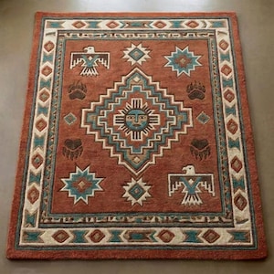 May include: A rectangular rug in a rust colour with a Southwestern design. It features geometric patterns, stylized eagles, bear paw prints, and a central sun face motif in turquoise, cream, and brown. The border has a repeating diamond pattern.