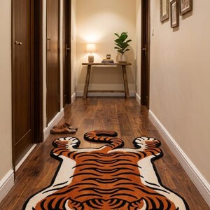 May include: A hallway runner rug shaped like a tiger, featuring orange and black stripes on a cream background. The rug has a detailed face with large eyes and a smiling mouth. The rug is placed on a wooden floor.