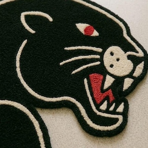 May include: Black panther rug with a cream-colored outline and details. The panther's eye is red, and its mouth is open, revealing white teeth and a red tongue. The rug has a textured, plush appearance.
