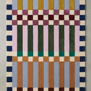 May include: A rectangular rug with a geometric pattern. The rug features alternating vertical stripes in shades of brown, blue, green, and pink, separated by checkered sections. The border is a navy blue and cream checkerboard design.
