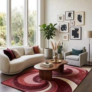 May include: A rectangular area rug featuring a swirling, concentric pattern in shades of deep burgundy, rose, and cream. The rug has a plush, textured pile and is positioned on a dark wood floor beneath a wooden coffee table in a living room.