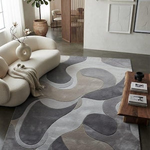 May include: A rectangular area rug featuring a multi-layered, abstract pattern of organic, flowing shapes in shades of charcoal gray, light gray, and muted beige. The rug has a textured, tufted surface with defined edges between the color blocks.
