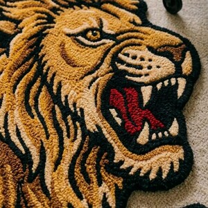Könnte beinhalten: Ein Teppich in Form eines brüllenden Löwenkopfes. Der Teppich hat ein detailliertes Design mit Farbtönen von Beige, Braun, Schwarz und Rot. Das Maul des Löwen ist geöffnet und zeigt weiße Zähne und eine rote Zunge. Der Teppich liegt auf einem hellen Teppich.