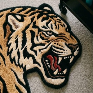 May include: A rug shaped like a tiger's head, featuring a detailed design with tan, black, and cream colors. The tiger's mouth is open, revealing red and white teeth. The rug is on a gray surface.