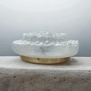 Pode incluir: Uma luminária de teto circular de montagem embutida com uma cúpula de vidro transparente espesso e texturizado, apresentando um padrão de superfície orgânico e borbulhante. A cúpula de vidro é montada numa base metálica circular simples com acabamento dourado fosco.