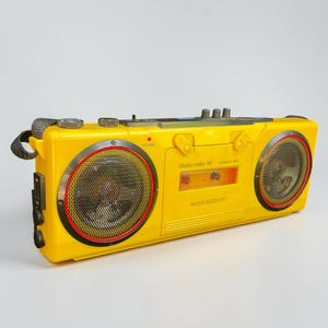 Puede incluir: Un radiocasete vintage de color amarillo brillante con dos altavoces, detalles en rojo y negro y un compartimento para cintas de cassette. El radiocasete tiene un asa y varios botones y diales. El texto "Saba radio Hi-Fi" es visible en la parte frontal.