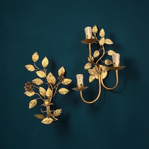 May include: Two gold-toned metal wall sconces featuring intricate leaf and rose motifs. One sconce holds a single candle, while the other holds two candles. Both fixtures have cream-colored candle covers with a faux-drip wax design.