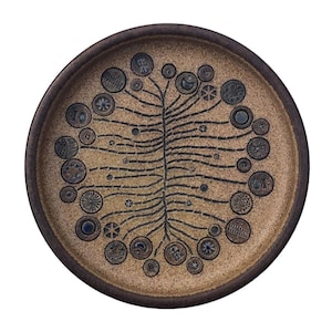 Puede incluir: Un plato redondo de cerámica marrón con una superficie texturizada. El plato presenta un diseño central de árbol estilizado con líneas radiales y patrones circulares y geométricos en marrón oscuro. El borde es de un tono marrón más oscuro.