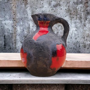 Puede incluir: Una jarra de cerámica hecha a mano con un exterior gris oscuro texturizado y acentos rojos vibrantes. La jarra presenta un asa curva y una forma orgánica única, perfecta para servir bebidas o como pieza decorativa.