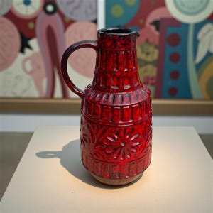 May include: A tall, red ceramic pitcher with a black rim and handle. The pitcher features a textured surface with floral and geometric patterns. The pitcher is displayed on a neutral surface, with artwork in the background.