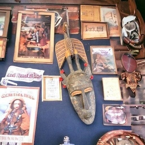Puede incluir: Una variedad de artículos vintage, incluyendo cuadros enmarcados, una máscara tribal y pequeños objetos decorativos, dispuestos sobre una superficie azul oscuro. La máscara es el punto focal, con una apariencia detallada y desgastada. Otros artículos incluyen un póster vintage y arte enmarcado pequeño.