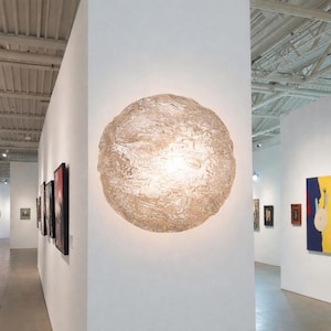 Könnte beinhalten: Eine große, strukturierte, kugelförmige Leuchte ist an einer weißen Wand in einer Kunstgalerie angebracht. Das Licht strahlt warm, und die Galeriewände zeigen gerahmte Kunstwerke. Die Oberfläche der Leuchte hat ein gesprenkeltes, organisches Aussehen.