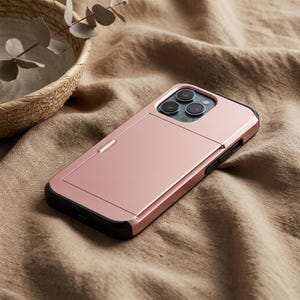 May include: A rose gold phone case with a black border. The case has a built-in card holder on the back and three camera lenses. The phone case is lying on a tan fabric surface.