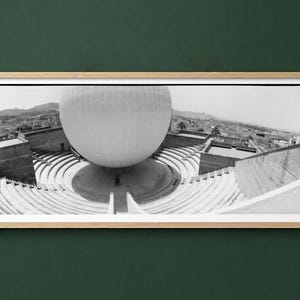 Può includere: Fotografia in bianco e nero di una grande struttura sferica sospesa sopra un anfiteatro circolare. L'immagine è incorniciata in una cornice di legno chiaro, su uno sfondo verde scuro. L'architettura è l'elemento principale.